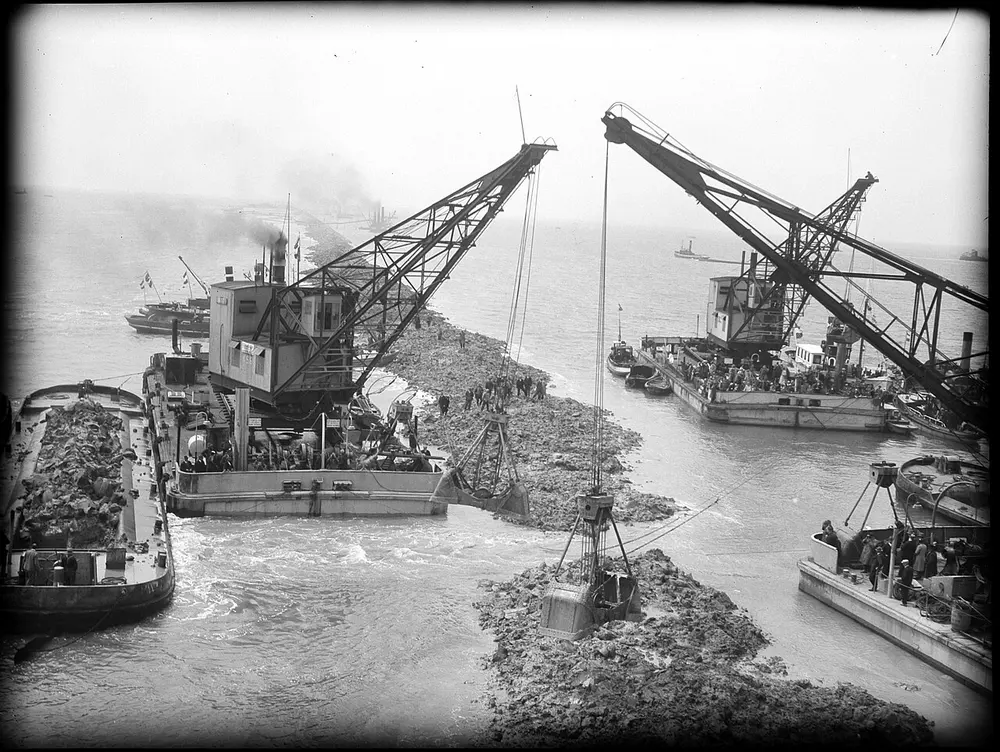 Zwart-witfoto uit 1932 van twee drijvende kranen met grijpers die materiaal storten in een smalle opening in een dijk in aanbouw, omringd door schepen en toeschouwers