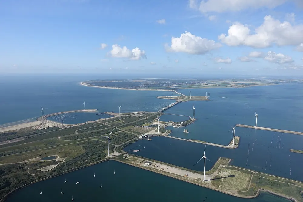 Luchtfoto van het eiland Neeltje Jans met windturbines, havens en natuurgebied, met op de achtergrond de rij pijlers van de Oosterscheldekering die over het water naar de overkant loopt