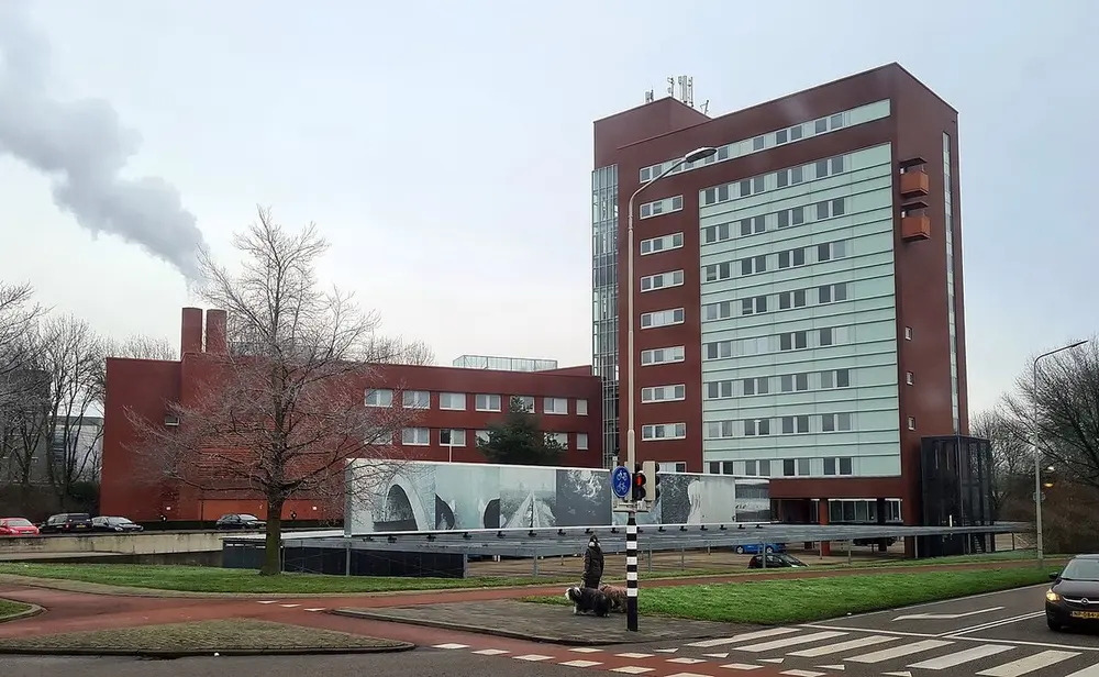 Kantoorgebouw van baksteen met een hoge toren met lichtgroene gevelpanelen, aan een kruispunt met zebrapad en fietspad, waar een vrouw met twee langharige honden loopt