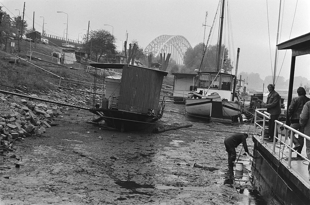 Zwart-witfoto van houten woonbootjes en een zeilboot die op de drooggevallen modderige rivierbodem liggen, met mannen op een steiger en op de achtergrond een boogbrug