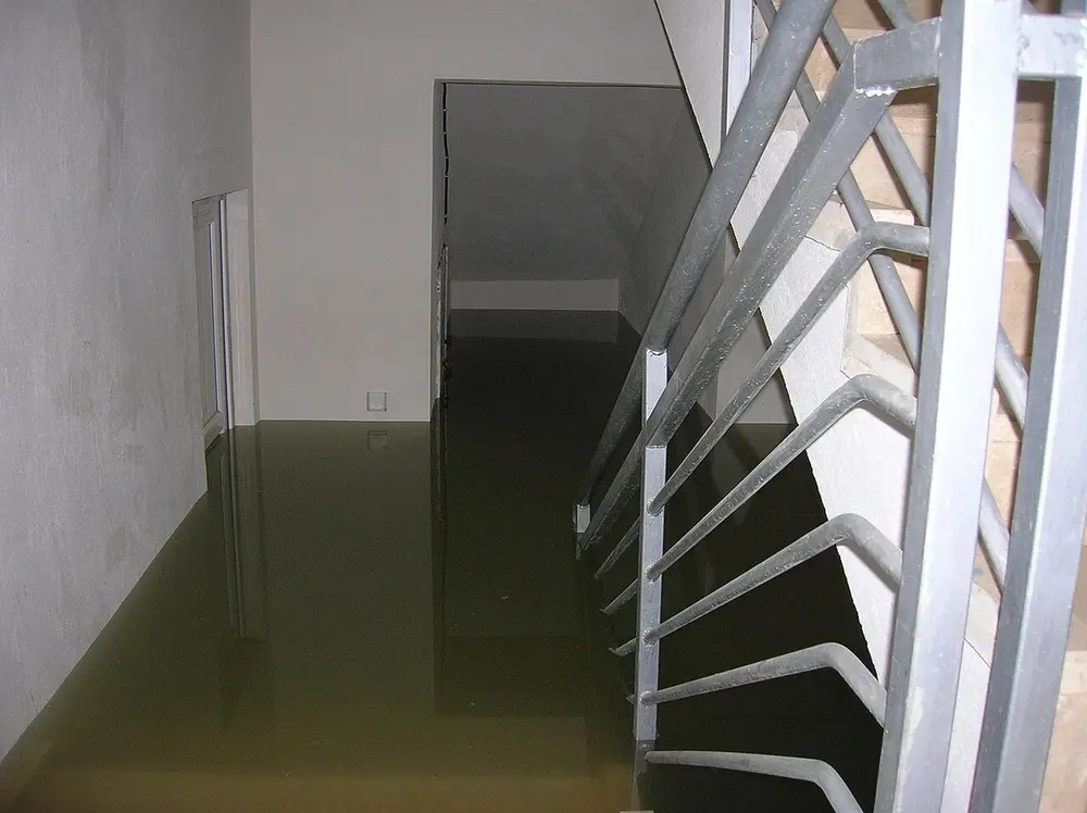 Kelder die tot boven de onderste traptreden onder water staat, gezien vanaf een metalen trap met leuning, met witte muren die in het troebele water weerspiegelen
