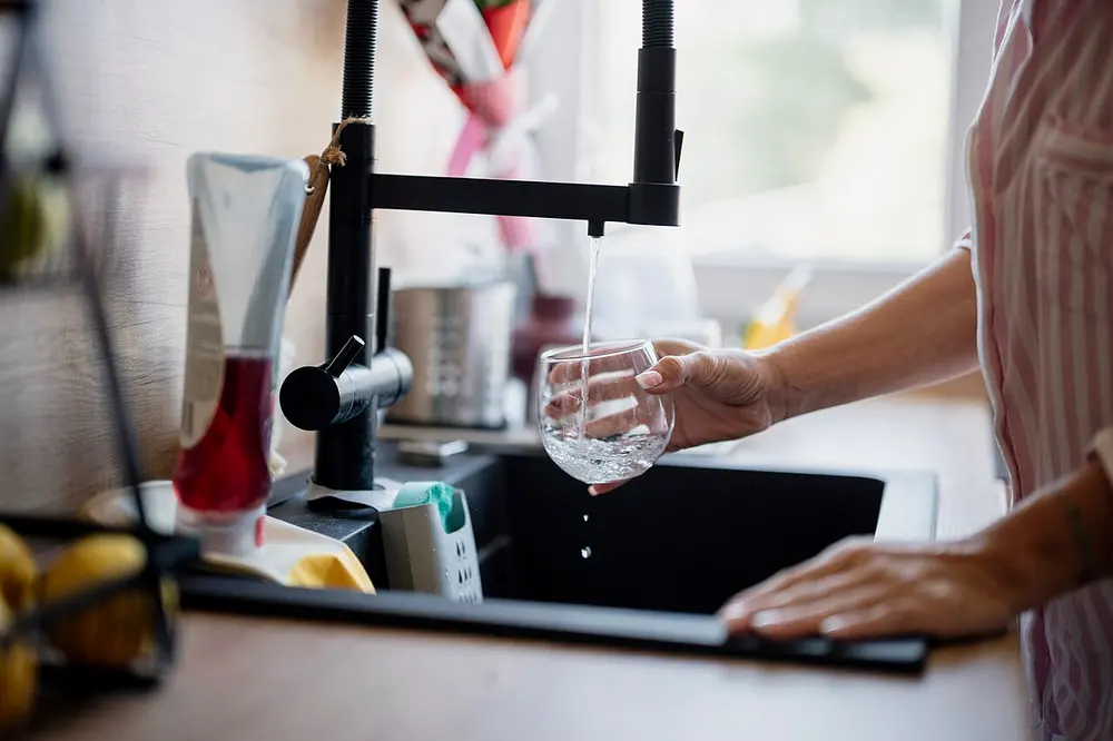 Iemand houdt een glas onder een zwarte keukenkraan waaruit een dunne straal water loopt, boven een zwarte spoelbak met een afwassponsje