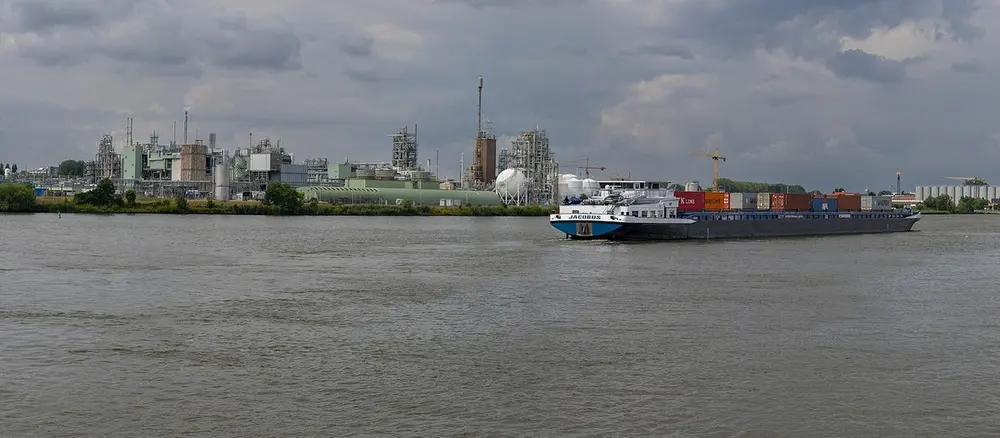Chemische fabriek met installaties en een witte bolvormige opslagtank aan de oever van een brede rivier, met op het water een binnenvaartschip met containers onder een bewolkte lucht