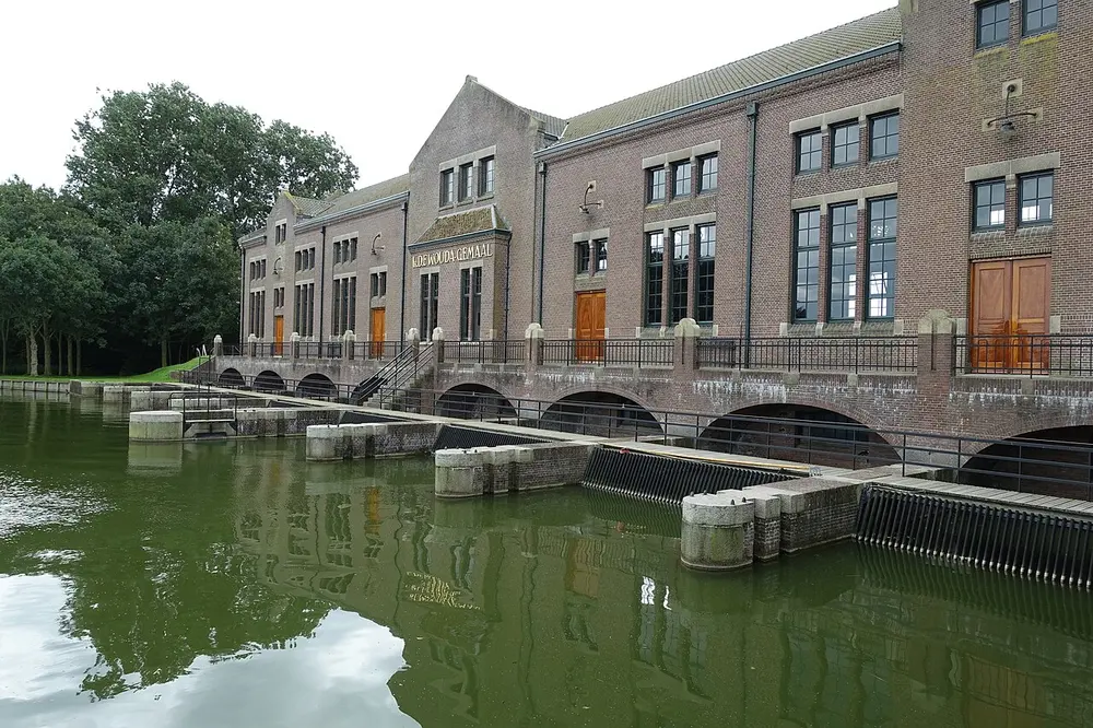 Bakstenen gemaalgebouw met de naam Ir. D.F. Wouda-gemaal op de gevel, grote ramen en houten deuren, met onder het gebouw boogvormige openingen aan groen water waarin de gevel weerspiegelt
