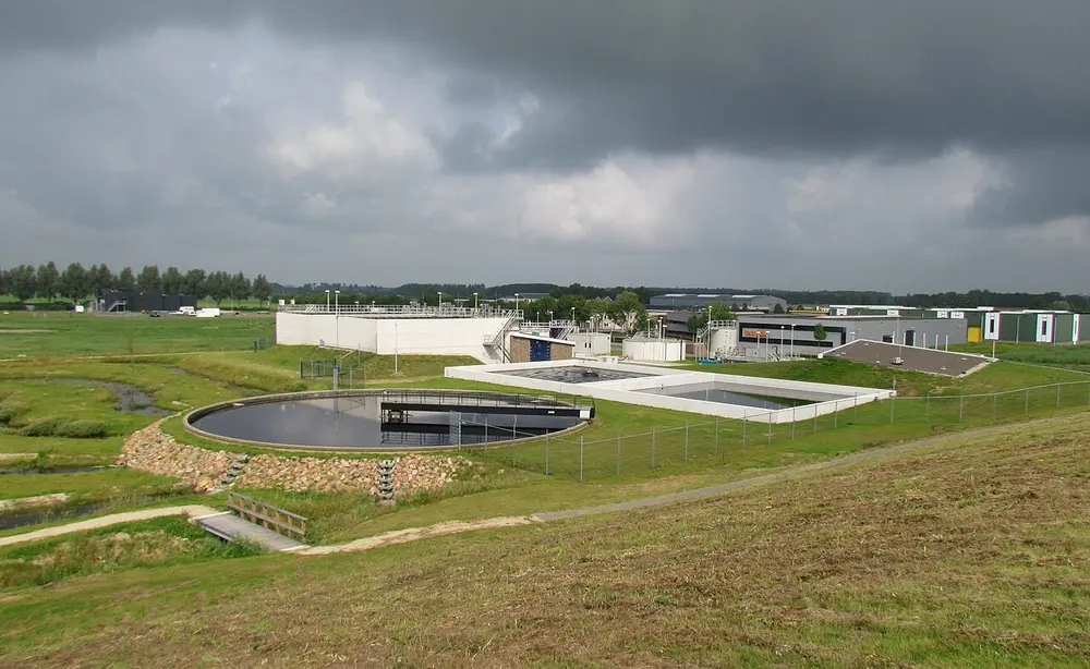 Rioolwaterzuivering in een vlak, groen landschap onder donkere wolken, met een ronde bezinktank op de voorgrond, rechthoekige betonnen bassins en witte gebouwen erachter