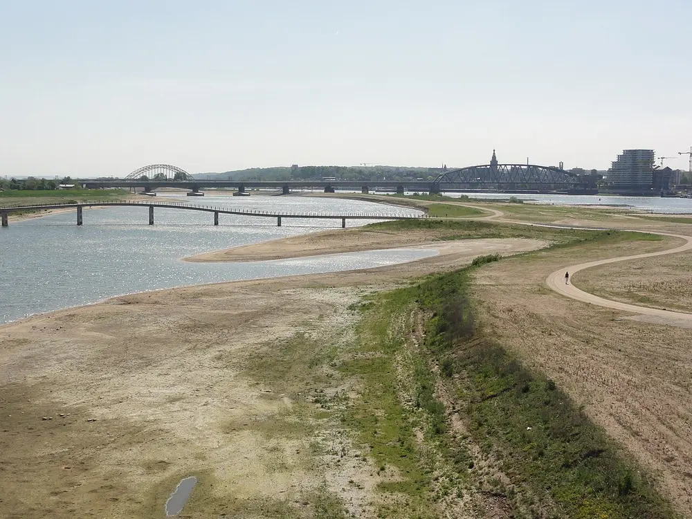 Brede rivierarm met zandige oevers en een kronkelend fietspad op de voorgrond, een lange lage voetbrug over het water en in de verte twee boogbruggen en een stad met een kerktoren