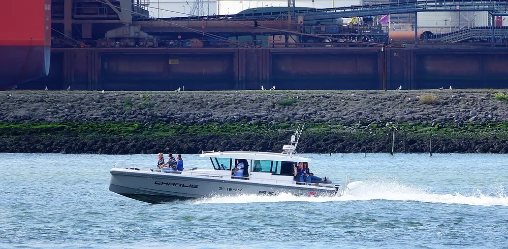 Grijze motorboot met open kuip vaart met hoge snelheid en een witte schuimstreep over de Nieuwe Waterweg, met enkele opvarenden aan boord en op de achtergrond een stenen oever en havenkade