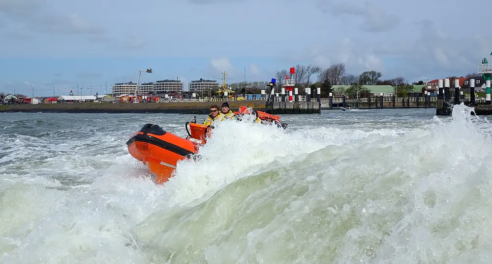 Oranje reddingboot van de KNRM met twee bemanningsleden in gele pakken stuitert over het schuimende hekgolfwater bij de havenmond van Hoek van Holland, met appartementengebouwen en havenbakens op de achtergrond