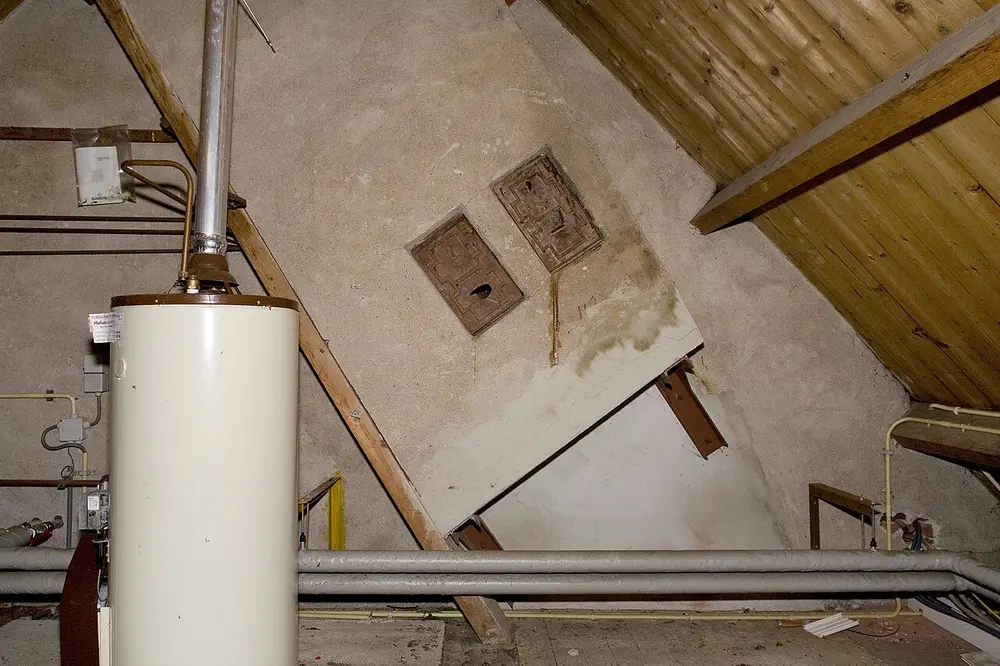 Crèmekleurige staande cilinder met een metalen afvoerpijp op een zolder onder een schuin houten dak, met geïsoleerde leidingen over de vloer