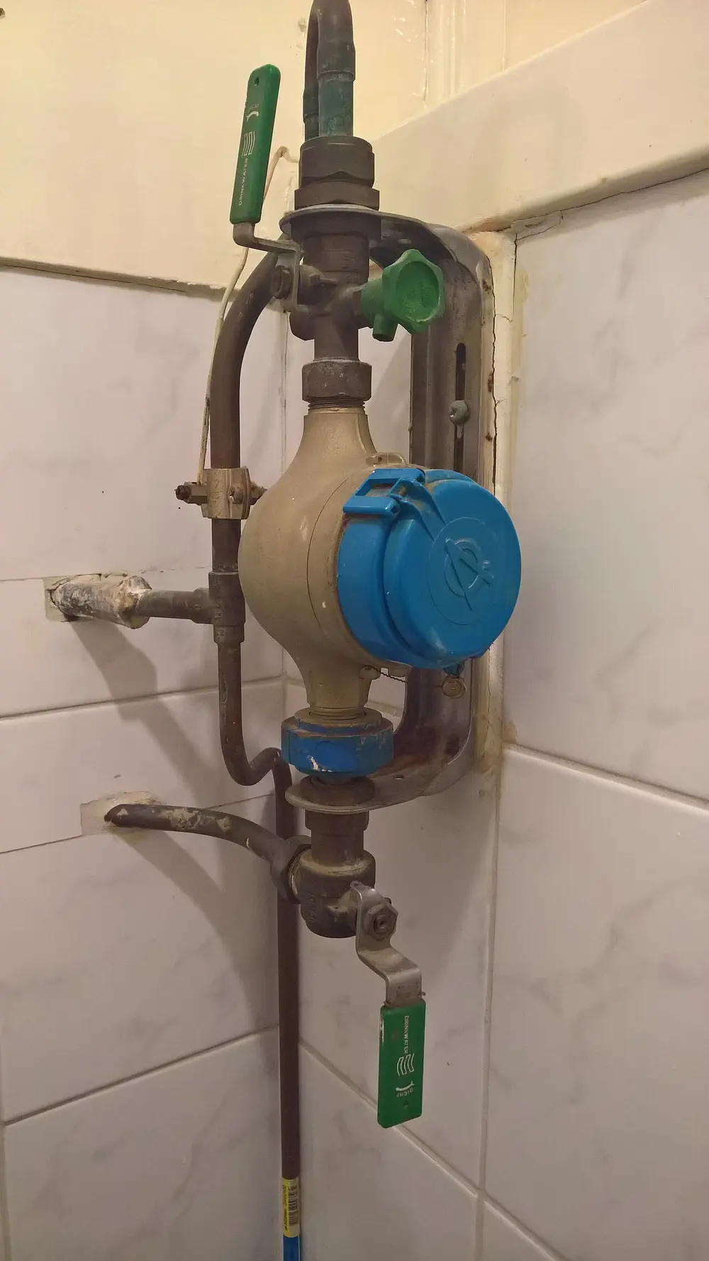 Watermeter met blauwe klepdeksel en groene afsluitkranen aan koperen leidingen, gemonteerd tegen een betegelde wand in een meterkast