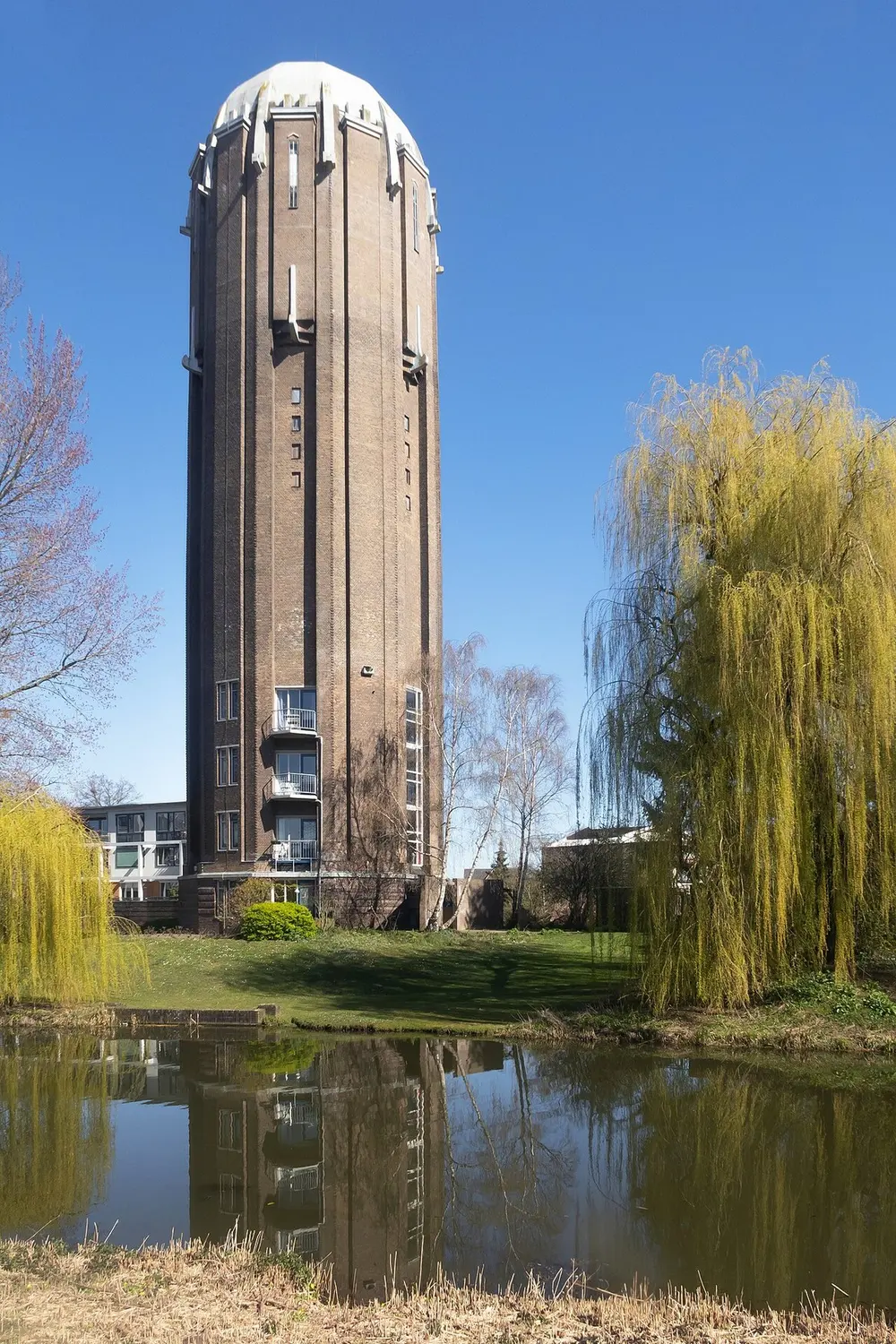 Hoge bakstenen watertoren met een lichte koepel en balkons in het onderste deel, gespiegeld in een vijver met treurwilgen op de voorgrond