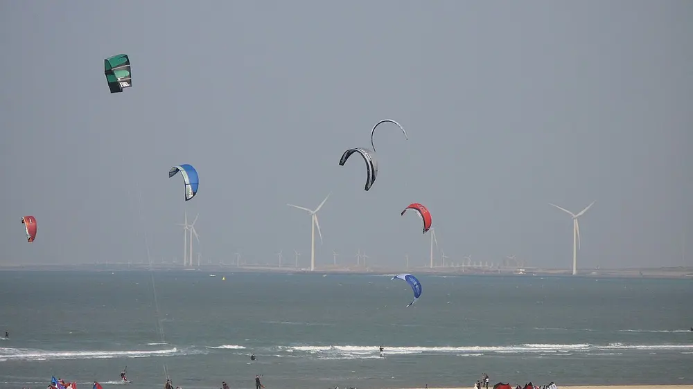 Zeven kleurige kites hoog boven zee met kitesurfers in de branding, een rij windturbines op een dam aan de horizon en op de voorgrond het strand met enkele mensen en opgezette kites