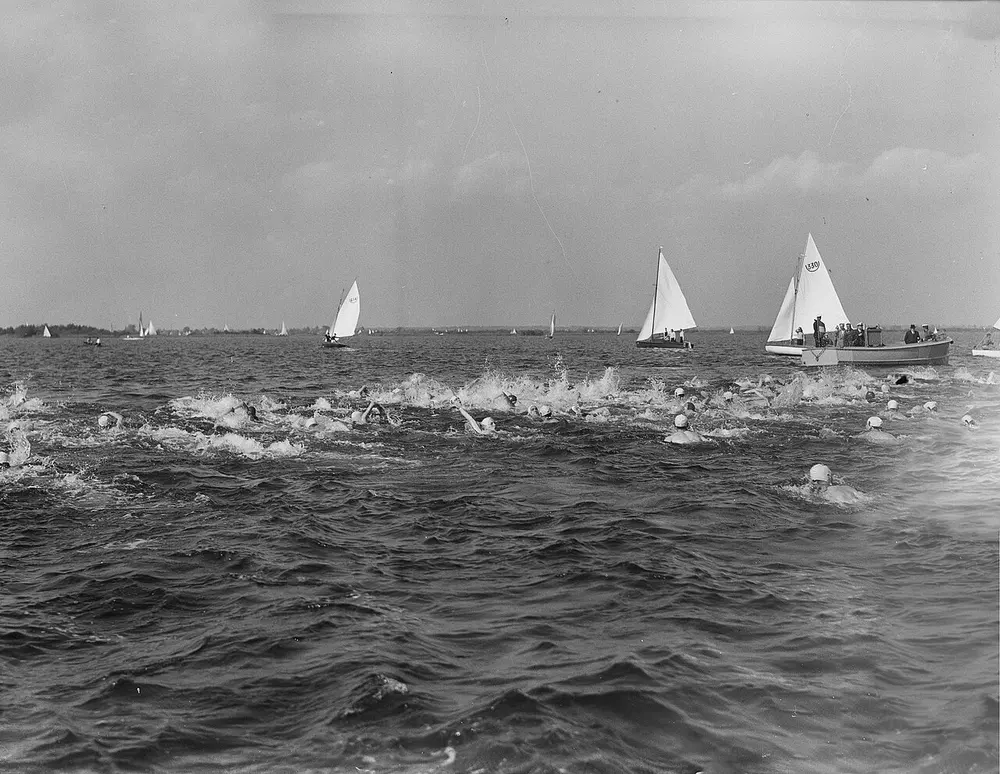 Zwart-witfoto uit 1950 van tientallen zwemmers met witte badmutsen die borstcrawlend door golvend water zwemmen, met zeilboten en een volgboot met toeschouwers op de achtergrond